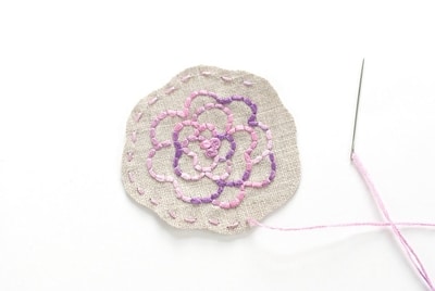 A hand-stitched purple flower design on a patch with the border still being stitched